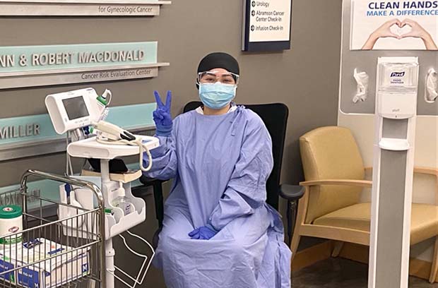 Nurse wearing PPE, making a peace sign, sitting by temperature check and hand sanitizing station 