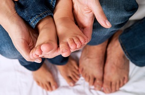 parents_holding_childs_feet_in_palm_of_hands_while_standing_barefoot_in_jeans_on_white_sheet
