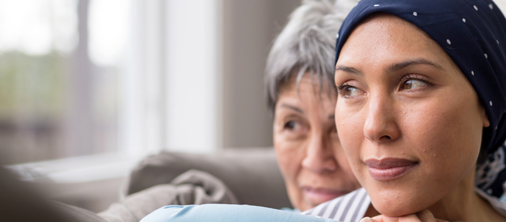 A mother and adult daughter are sitting on the couch looking out a window. The mother hugs her daughter, who is wearing a head covering.