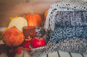 A still-life featuring an apple, pomegranate and two small pumpkins surrounding a votive candle. All items are positioned next two four knitted blankets stacked on top of each other.
