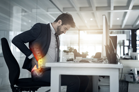 man in suit sits at desk in front of a computer and holds his aching back.