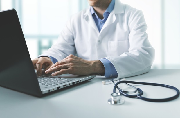 A stethoscope is next to a male doctor who is typing on a laptop