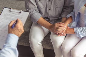 Couple sit next to each other on examining table holding hands as a doctor sees them and takes their information on a clipboard