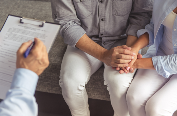 Couple sit next to each other on examining table holding hands as a doctor sees them and takes their information on a clipboard